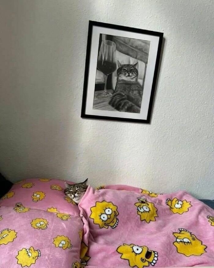 Cat tucked in bed under a pink Lisa Simpson blanket with a framed cat portrait on the wall showing animal shenanigans.
